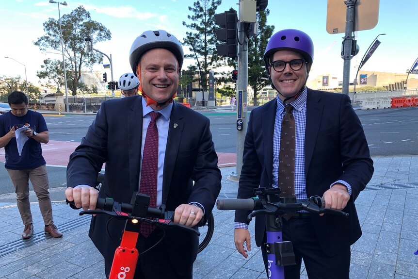 Two men wearing helmets smiling while stationary on e-scooters