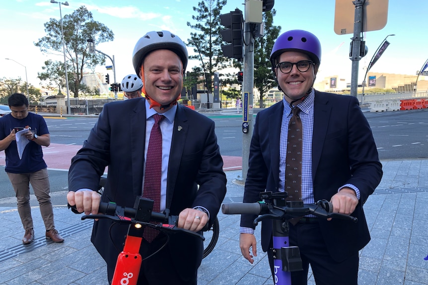 Two men wearing helmets smiling while holding e-scooters.