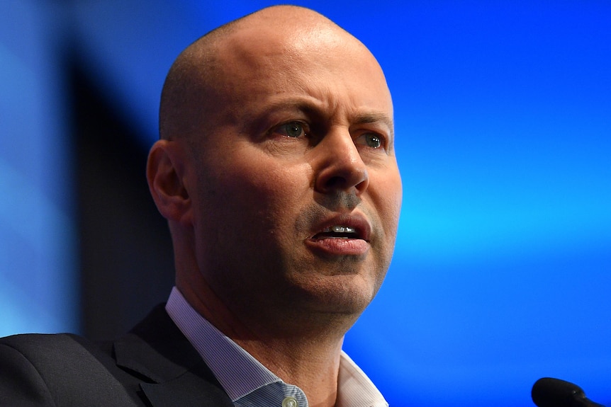 Josh Frydenberg has a neutral but determined expression as he speaks at a lectern