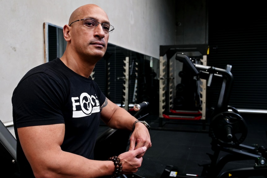 a muscular man leans against gym equipment and looks into the camera. Machines in a warehouse-style gym are behind him