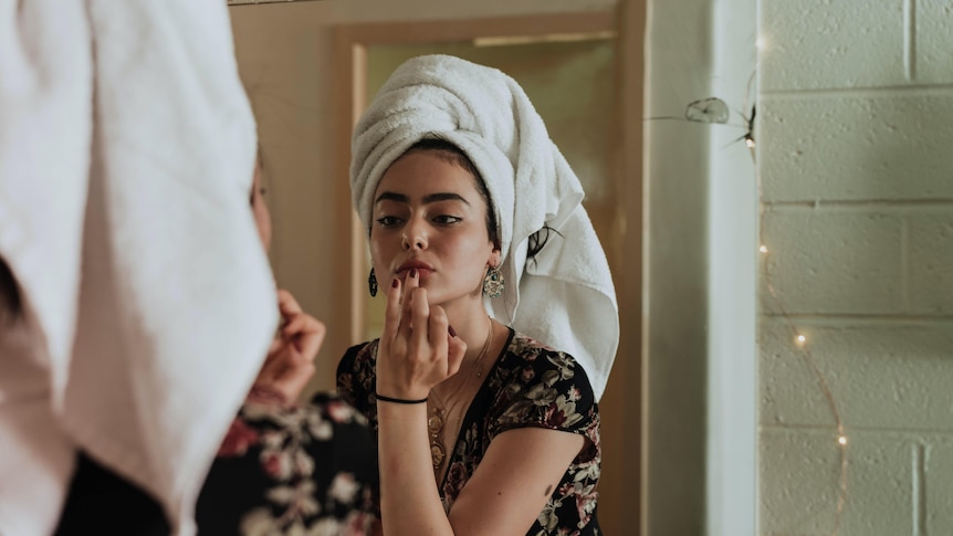 A woman with a towel on her head looks at her face in a mirror. 