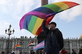 A white man in a cap waves a rainbow flag at a rally in front of historic Russian buildings.