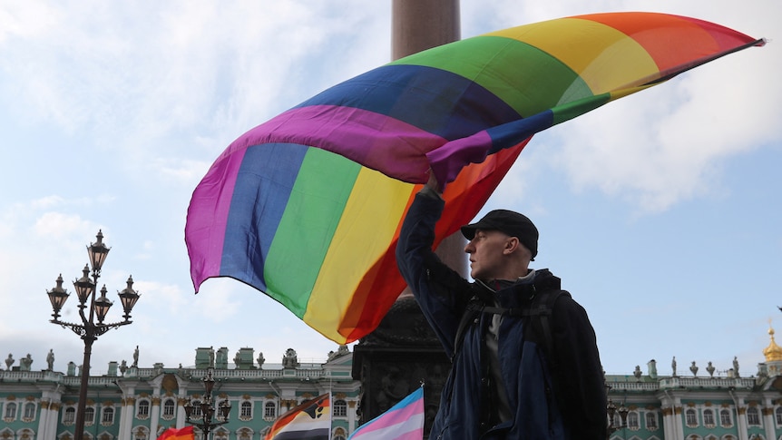 A white man in a cap waves a rainbow flag at a rally in front of historic Russian buildings.
