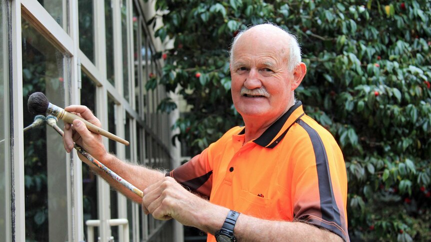 Older man in bright orange tshirt holding a paint brush and balancing rod against a glass wall