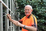 Older man in bright orange tshirt holding a paint brush and balancing rod against a glass wall