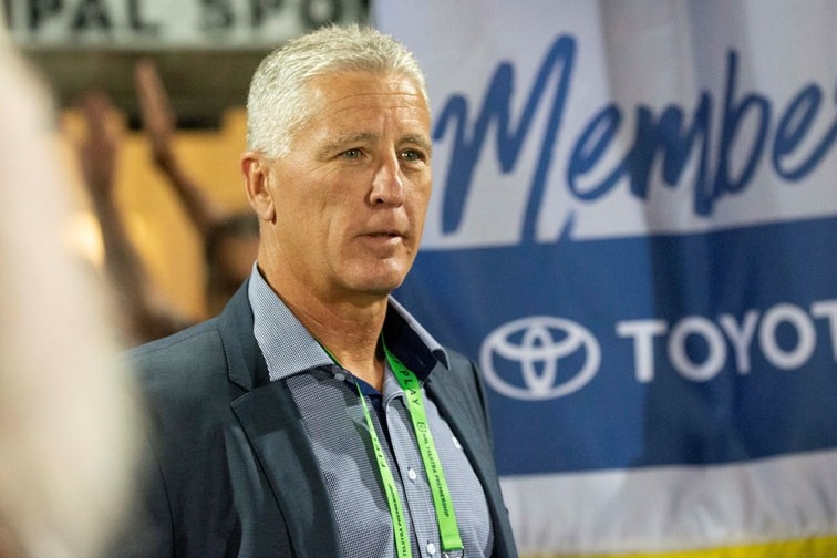 Man with short grey hair in blue suit jacket and shirt wearing lanyard standing in front of large sign