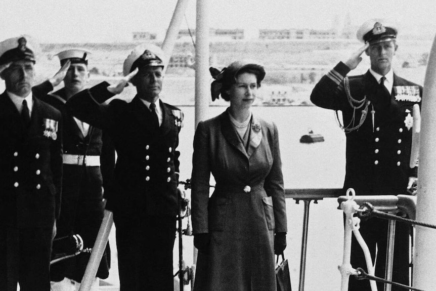 A black and white photograph of Princess Elizabeth on a naval ship with Prince Philip saluting in the background.