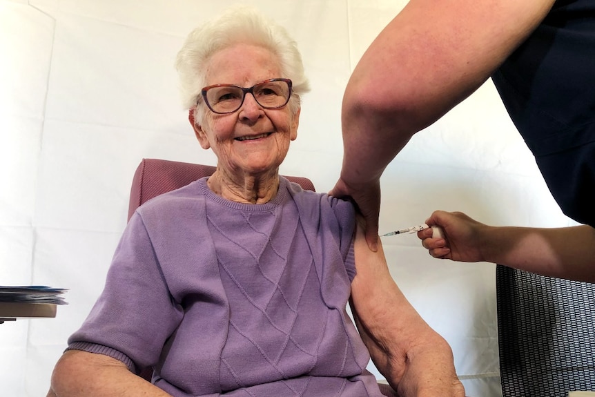 An elderly woman wearing glasses smiles as she has a vaccination