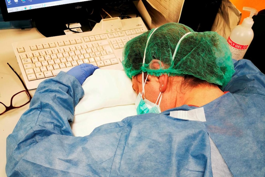 A woman in scrubs and a face mask sleeps at her desk