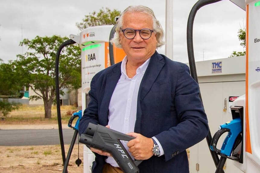 A man with grey hair and wearing glasses and a blue suit jacket stands at an electric vehicle charging station.