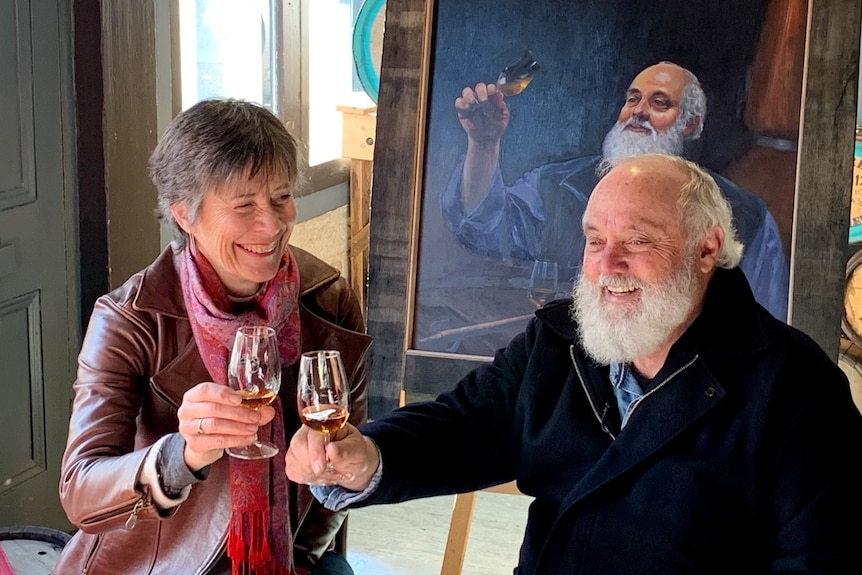 A middle-aged woman and man sit on a barrel drinking whisky in front of a portrait of the man