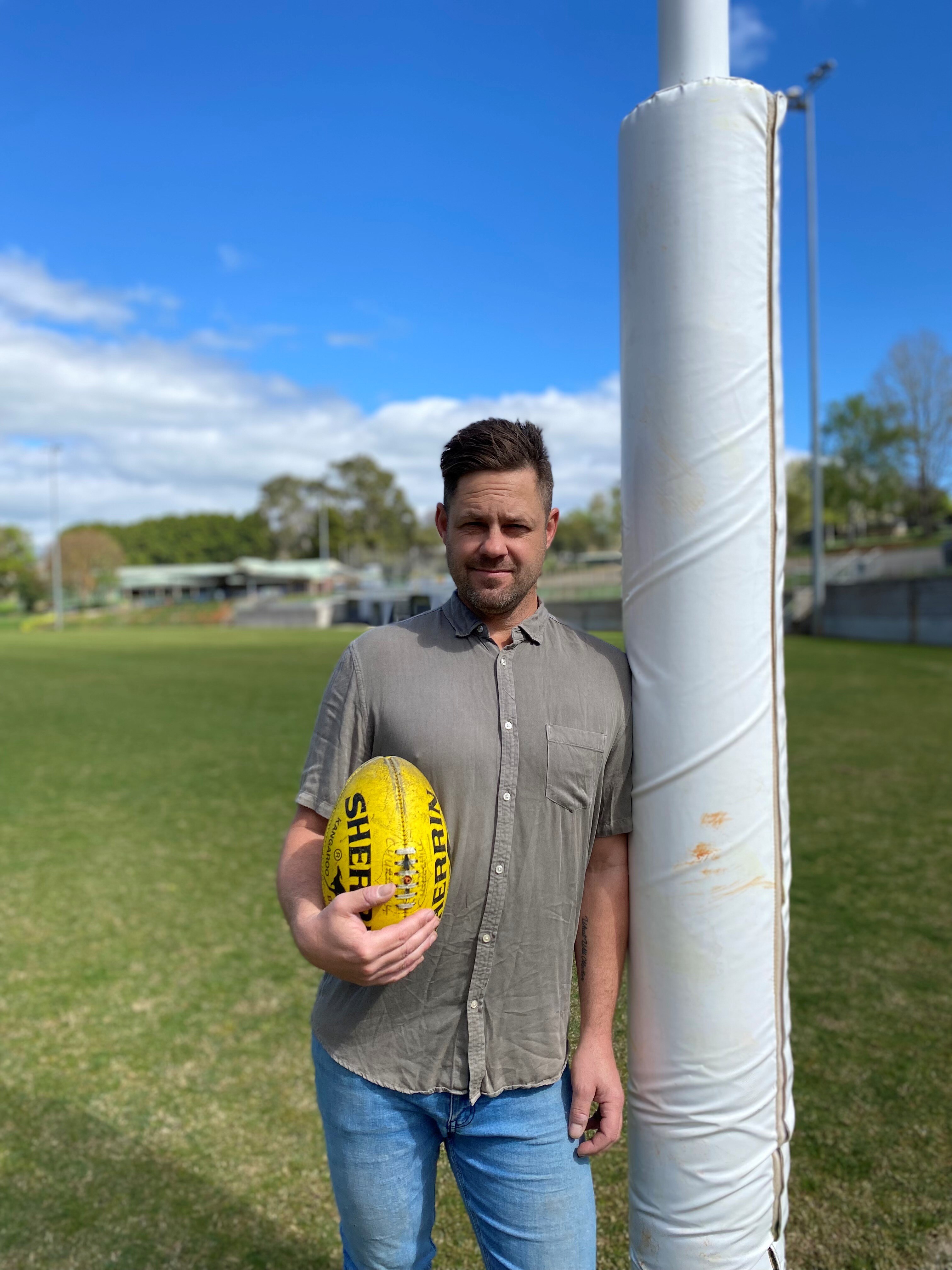 A middle-aged man with short brown hair leaning against an AFL post, while holding a yellow football.