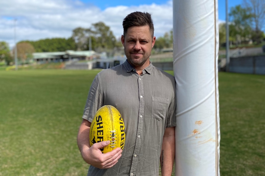 A middle-aged man with short brown hair leaning against an AFL post, while holding a yellow football.