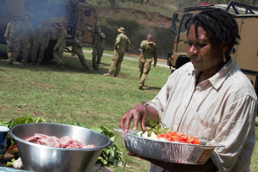 A woman with hair in short black tight braids handles cut vegetables in a large dish with army troops standing in the background