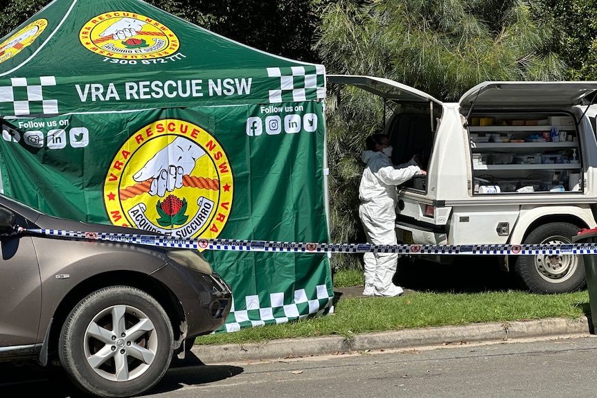 A green gazebo shields the view of a house and a person in white protective clothing stands at a vehicle.