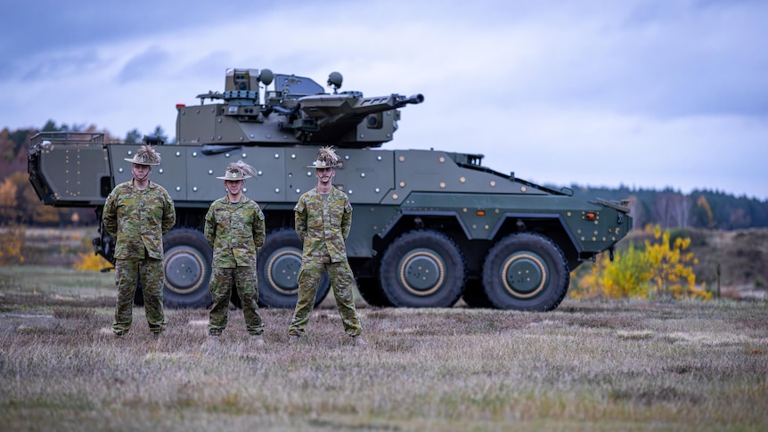 Three men in military uniform stand in front of an armoured vehicle in a field.