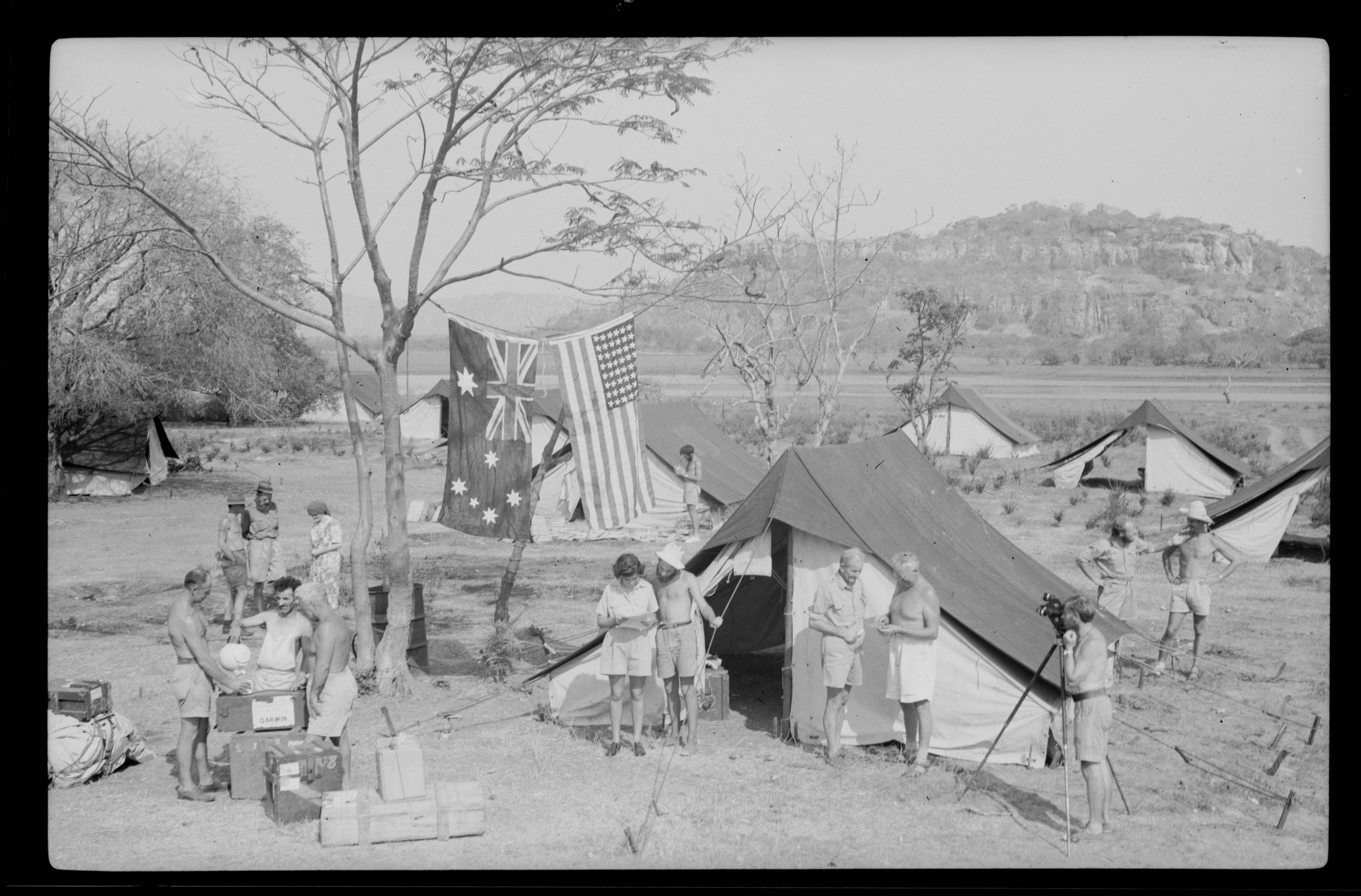A black and white photo of tents in the outback with Australian and American flags hanging from a tree
