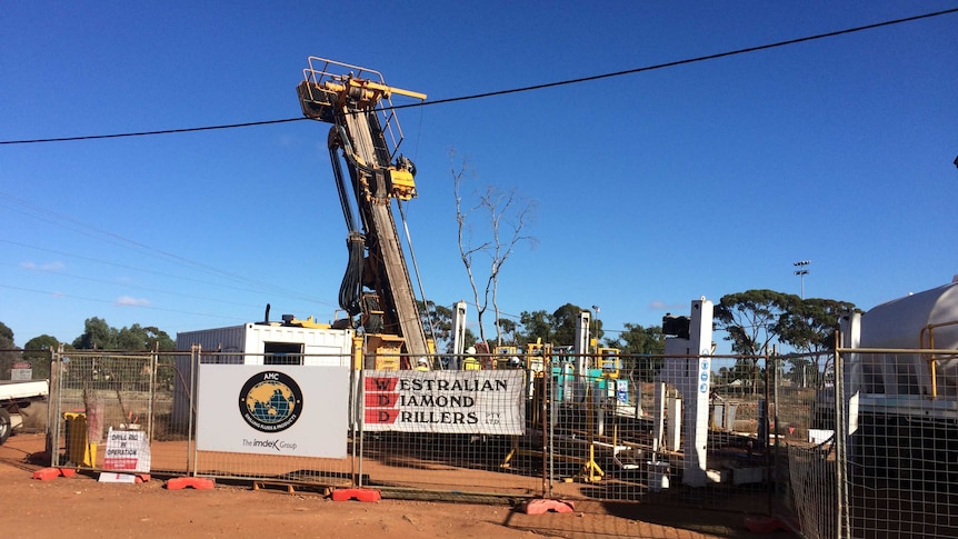 A weld mesh fence surrounds a large, vertical diamond drill machine, water truck and small site office in suburban Kalgoorlie