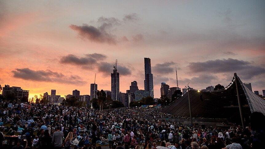 A crowd congregates in an outdoor space in front of a public stage at sunset. The Melbourne skyline is in the background.