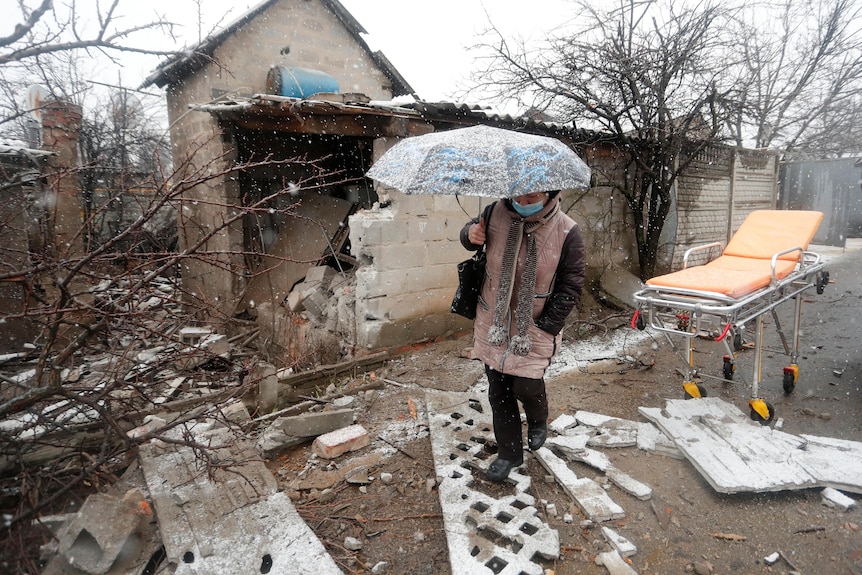 A woman in winter clothes and holding an umbrella walks next to a damaged house, amid light snow.