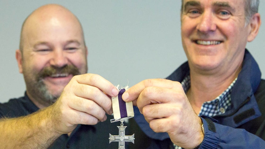 Two smiling men hold up a cross-shaped war medal.