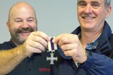 Two smiling men hold up a cross-shaped war medal.