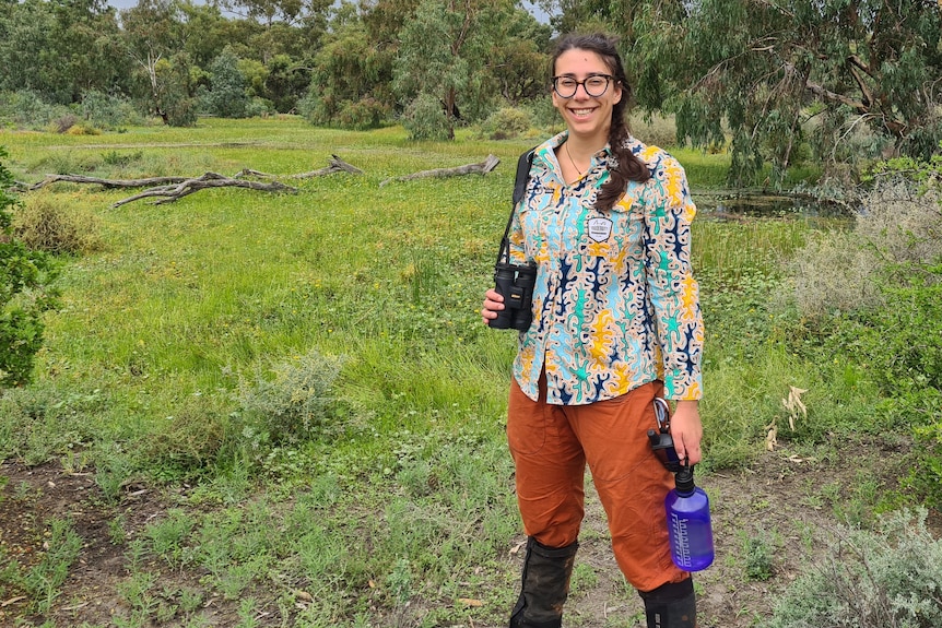 A woman with long hair in a pony tail with glasses holds a water bottle and binoculars in her hands surrounded by grass. 