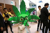 A person stands wearing a marijuana leaf-shaped mascot head and scientist's lab coat.