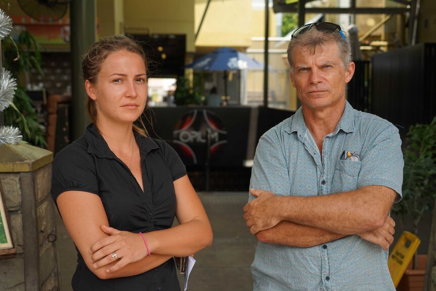 A young woman and her father stand with arms crossed outside a bar in daylight.