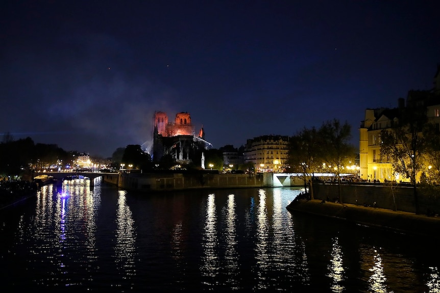 A wide shot shows a burning cathedral on an island in a river with street lights reflecting across the surface of the water