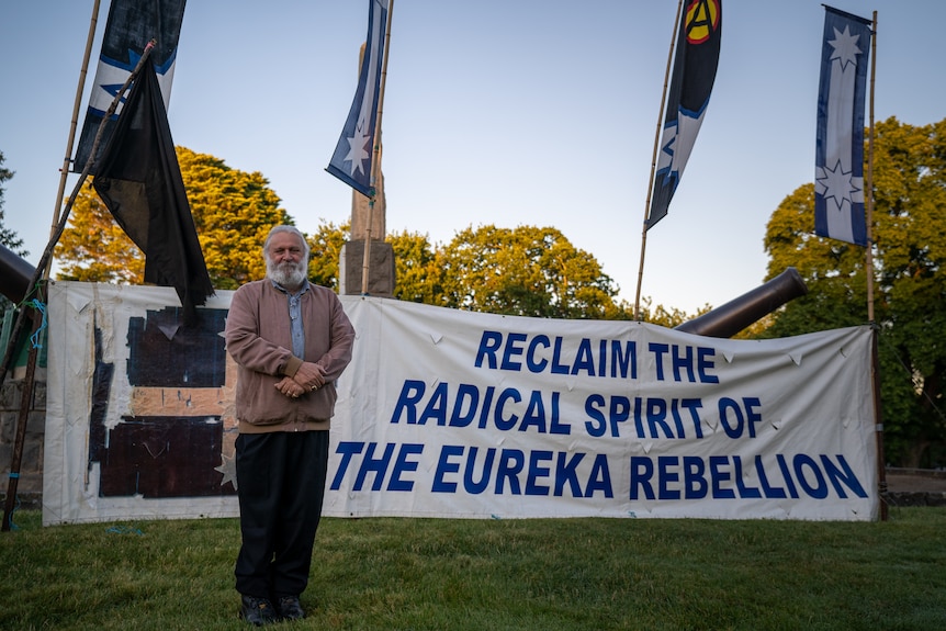 An older man with a long white beard stands in front of a large banner that urges the reclamation of the Eureka spirit.