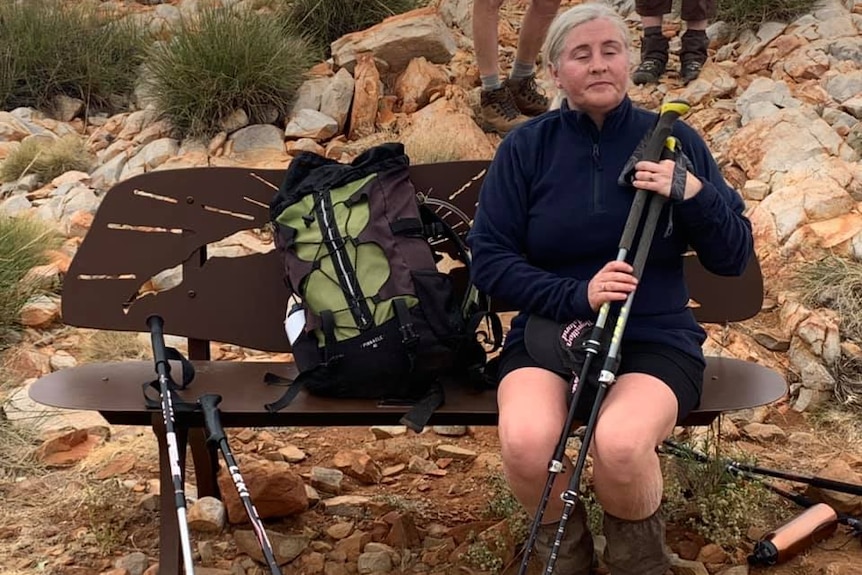 Woman holding hiking poles sits on a bench on a rocky trail.