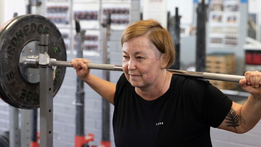 A middle-aged woman has weights on her shoulders and prepares to lift.
