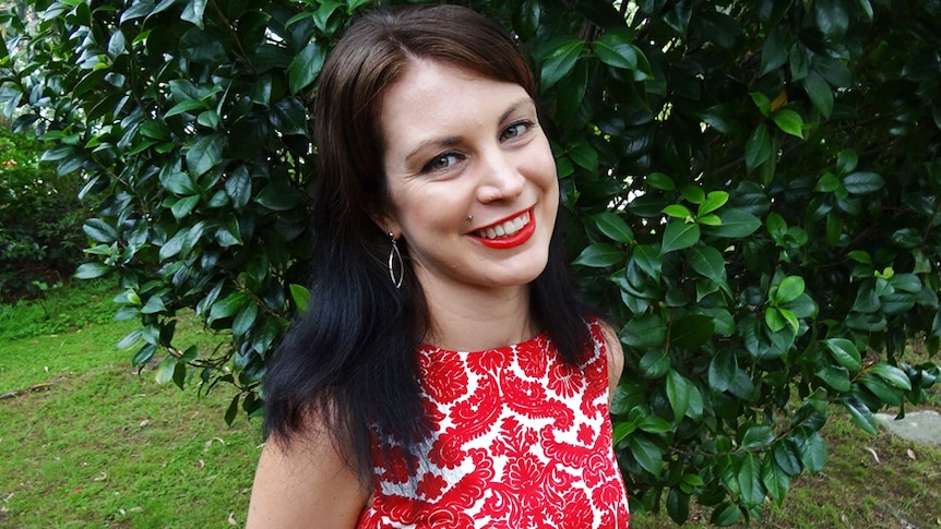 A smiling, dark-haired woman in a red and white dress stands in a backyard in front of a tree with lots of dark green leaves.