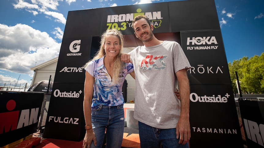 A young woman with blonde hair stands next to a young man with a beard in front of an ironman sign