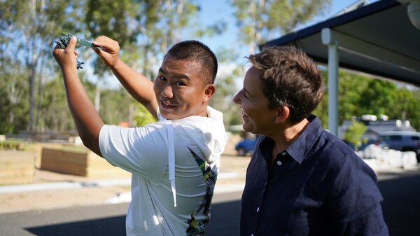 Young man wearing t-shirt standing outside holding a fresh tiger prawn in the air, a woman looks on smiling.