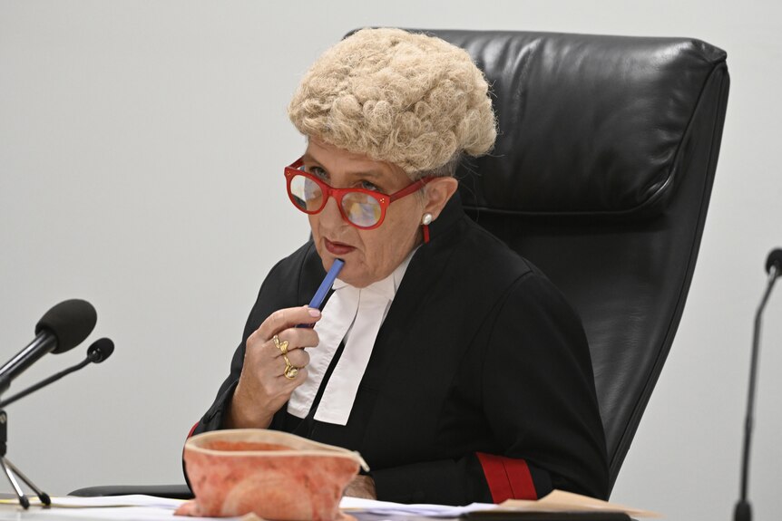A female judge wearing a white wig and red glasses in court