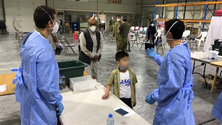 People wearing masks walk toward medical professionals wearing blue gowns and masks in a large hangar space