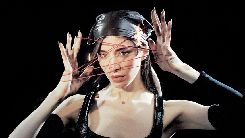 Caroline Polachek with black background holding her hands in front of her face with elastic between her fingers.