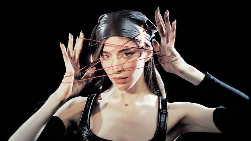 Caroline Polachek with black background holding her hands in front of her face with elastic between her fingers.