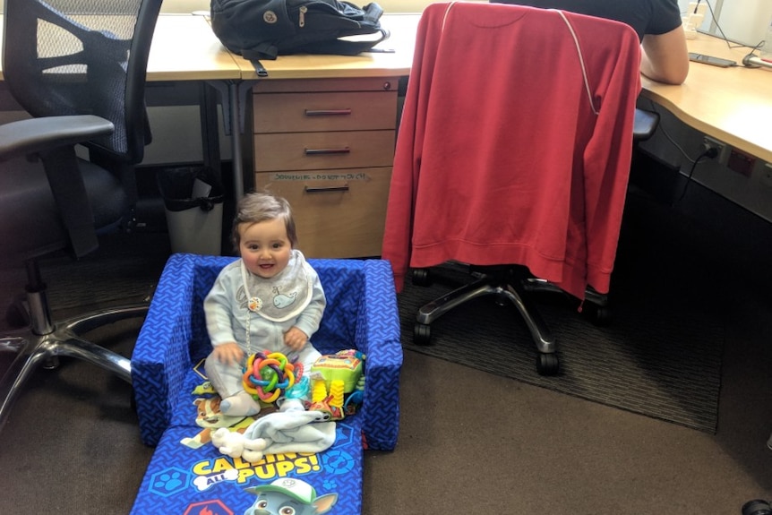 a baby sits on the floor of an office. There is a man sitting at his desk in the background.