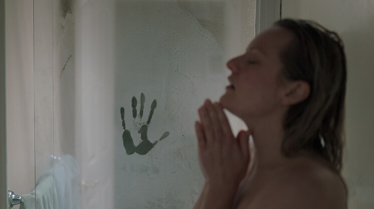 A woman showers with eyes closed, next to her on misty shower door is a large hand print.