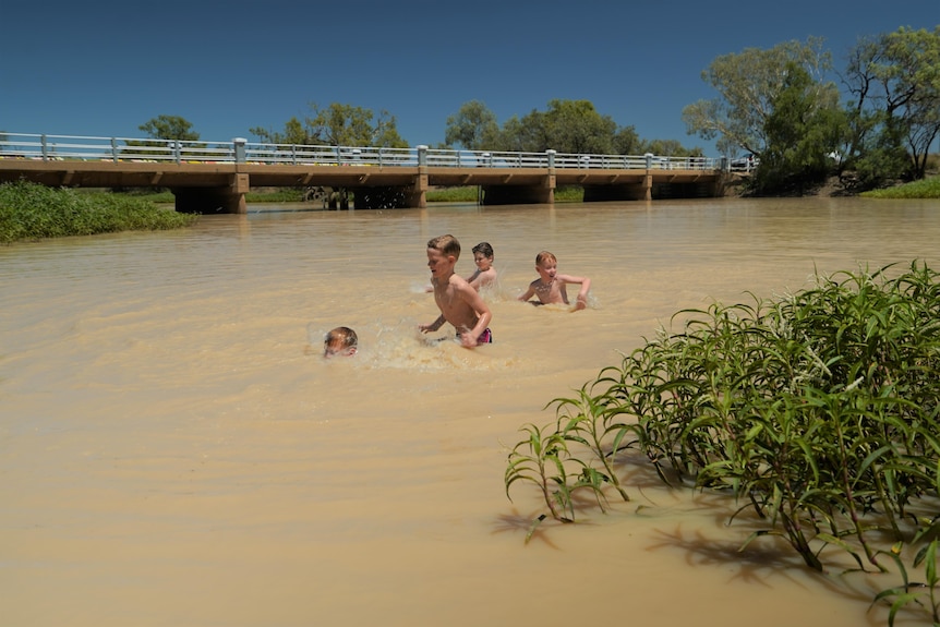 Four young boys swim in a brown river next to a bridge 