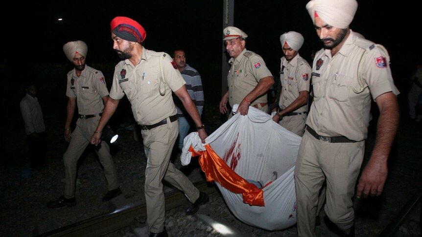 Men wearing turbans and dressed in khaki uniforms carry something heavy in a white sheet with dark red stains