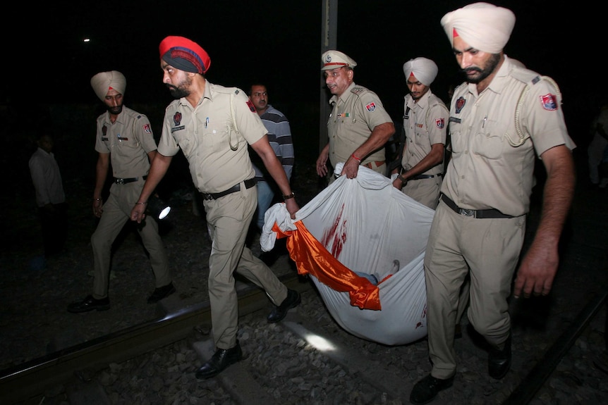 Men wearing turbans and dressed in khaki uniforms carry something heavy in a white sheet with dark red stains
