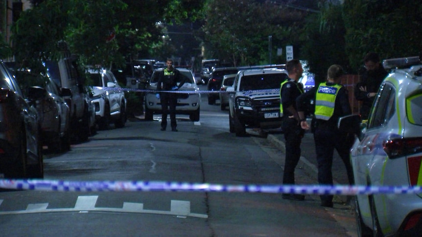 Police officers stand on a dark inner-city street at night, which is taped off by police tape.