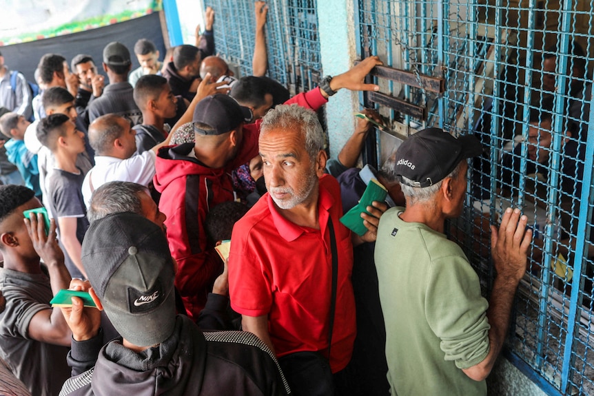 Dozens of men stand in front of a window with metal netting over it, reaching their hands to it