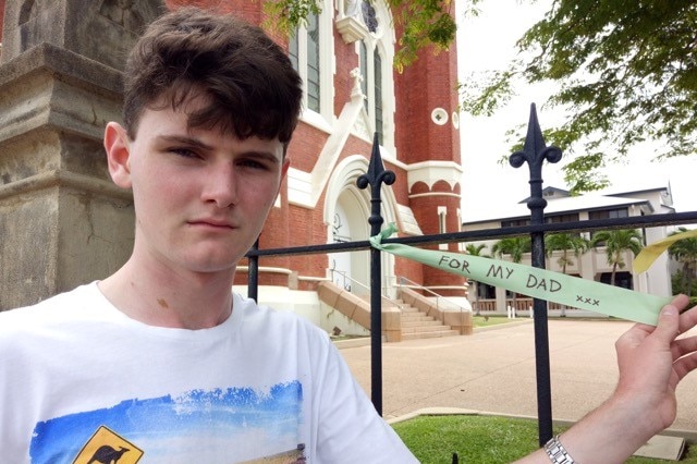 A teenage boy's arm holds a ribbon tied onto a cathedral fence which reads "for my dad"