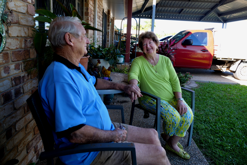 a man and woman sit on their front porch at a table and chairs, looking at each other and smiling 