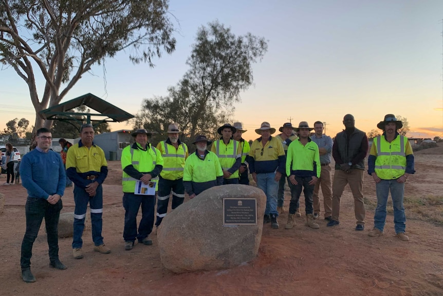 A group of men in high-vis gear stand around a rock with a plaque on it. It is dusk in the desert.